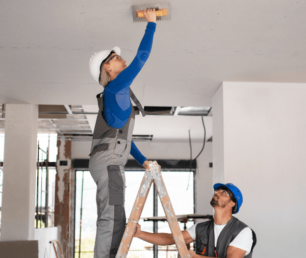 Two construction workers, one standing on a ladder and another assisting, installing or inspecting equipment on a ceiling in a building under renovation. both are wearing hard hats and safety gear. Two construction workers, one standing on a ladder and another assisting, installing or inspecting equipment on a ceiling in a building under renovation. both are wearing hard hats and safety gear.