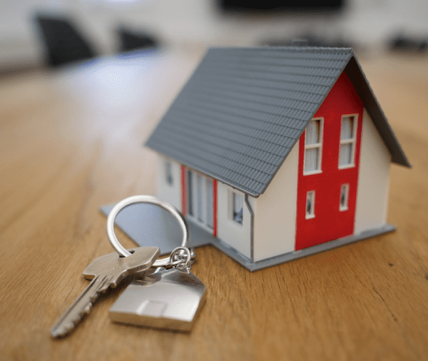 A small model house with red walls and a gray roof, accompanied by keys with a keychain, placed on a wooden table, suggesting themes of real estate or homeownership. A small model house with red walls and a gray roof, accompanied by keys with a keychain, placed on a wooden table, suggesting themes of real estate or homeownership.