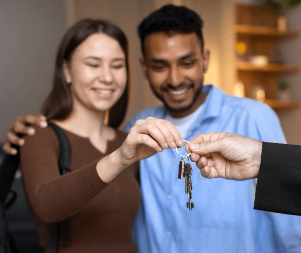 A smiling young couple receives a set of house keys from a person's hand, foreground focused on keys, with a cozy room setting in the background. A smiling young couple receives a set of house keys from a person's hand, foreground focused on keys, with a cozy room setting in the background.