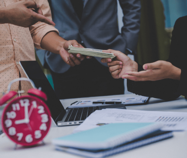 Person handing over a stack of cash to another person across a table with documents and a laptop; a red alarm clock is also visible. Person handing over a stack of cash to another person across a table with documents and a laptop; a red alarm clock is also visible.